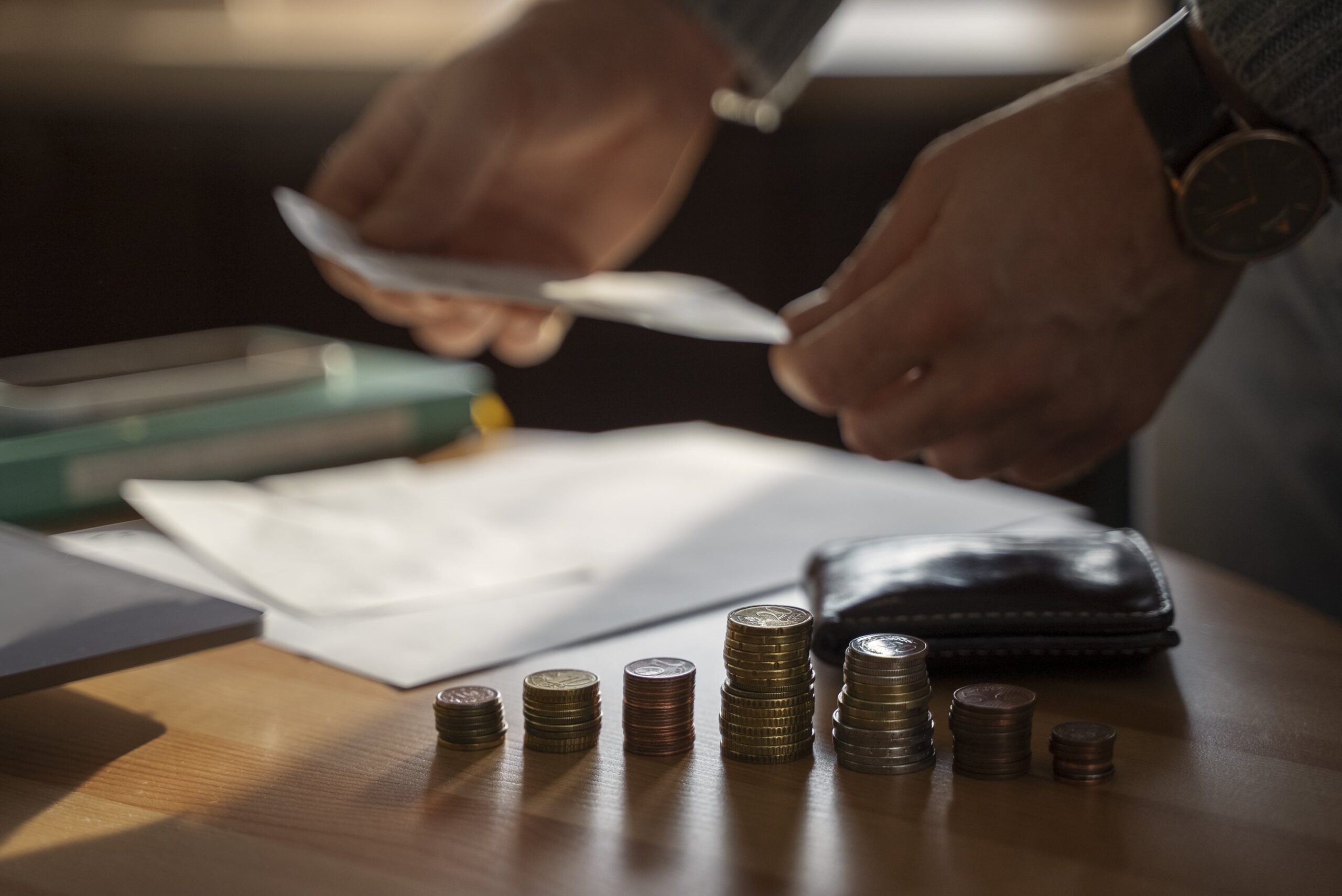 A person organizes coins in stacks on a wooden table while holding papers, suggesting financial planning or budgeting.