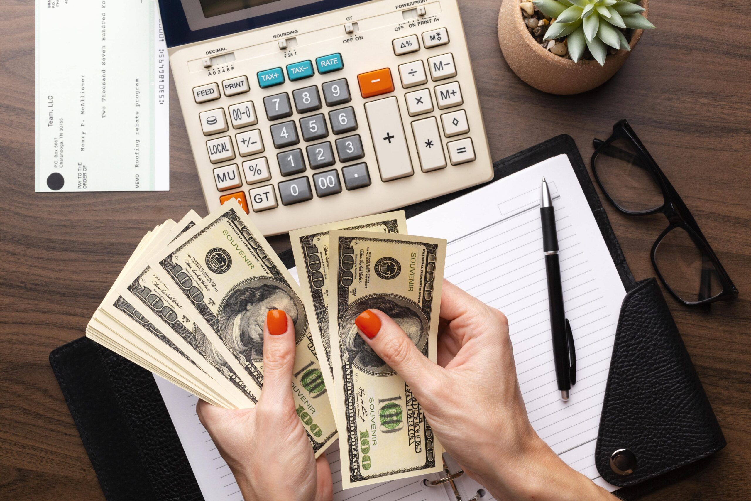 Hands holding stacks of hundred-dollar bills with a calculator, notebook, and glasses on a wooden desk, indicating financial planning or budgeting.