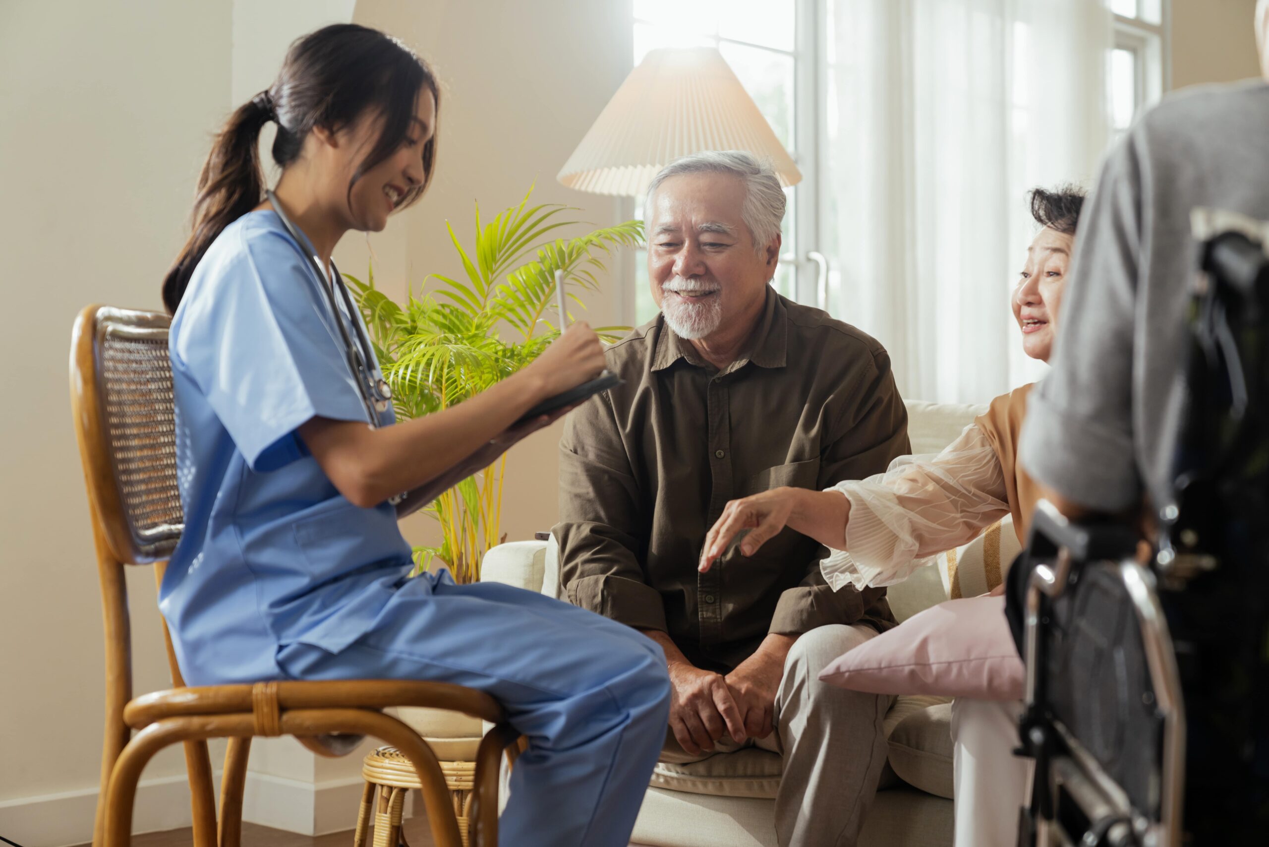 A healthcare worker in scrubs engages with an elderly couple, sharing a lighthearted moment. The scene highlights compassionate care in a home setting.