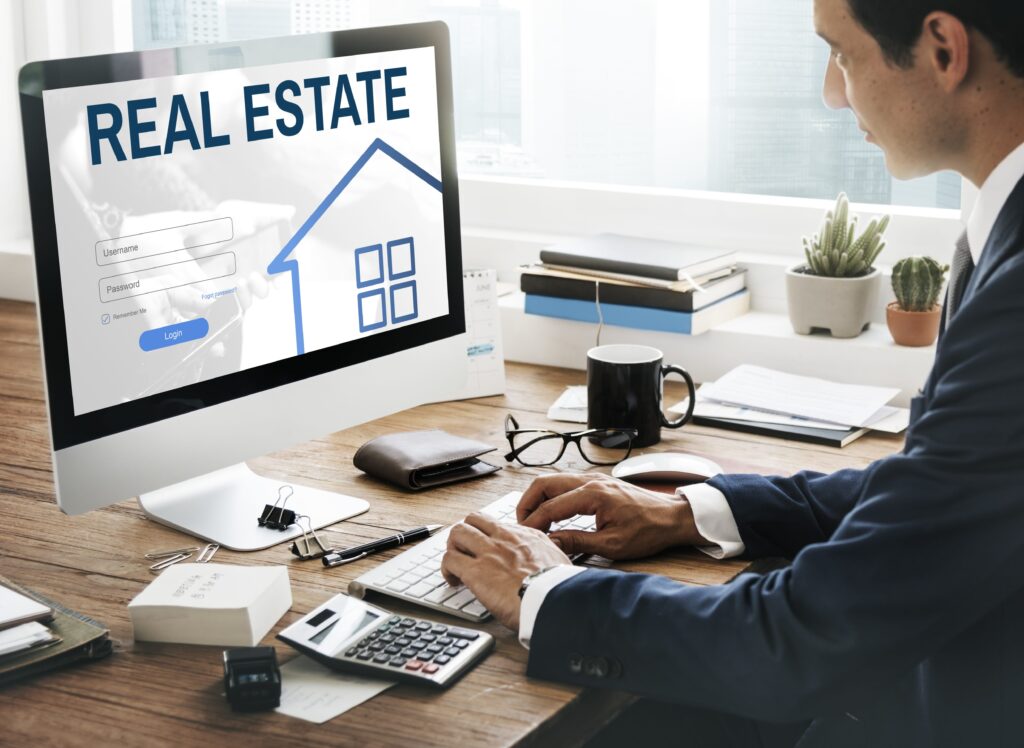 A businessman in a suit is logging into a real estate platform on a computer. The workspace features stationery, a calculator, and plants, emphasizing a professional setting.