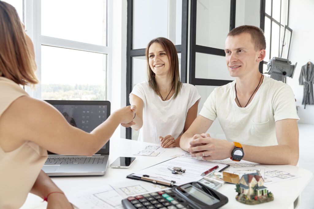 A woman shakes hands with a smiling client in a bright office, discussing a project. Papers, a laptop, and a miniature house are on the table.