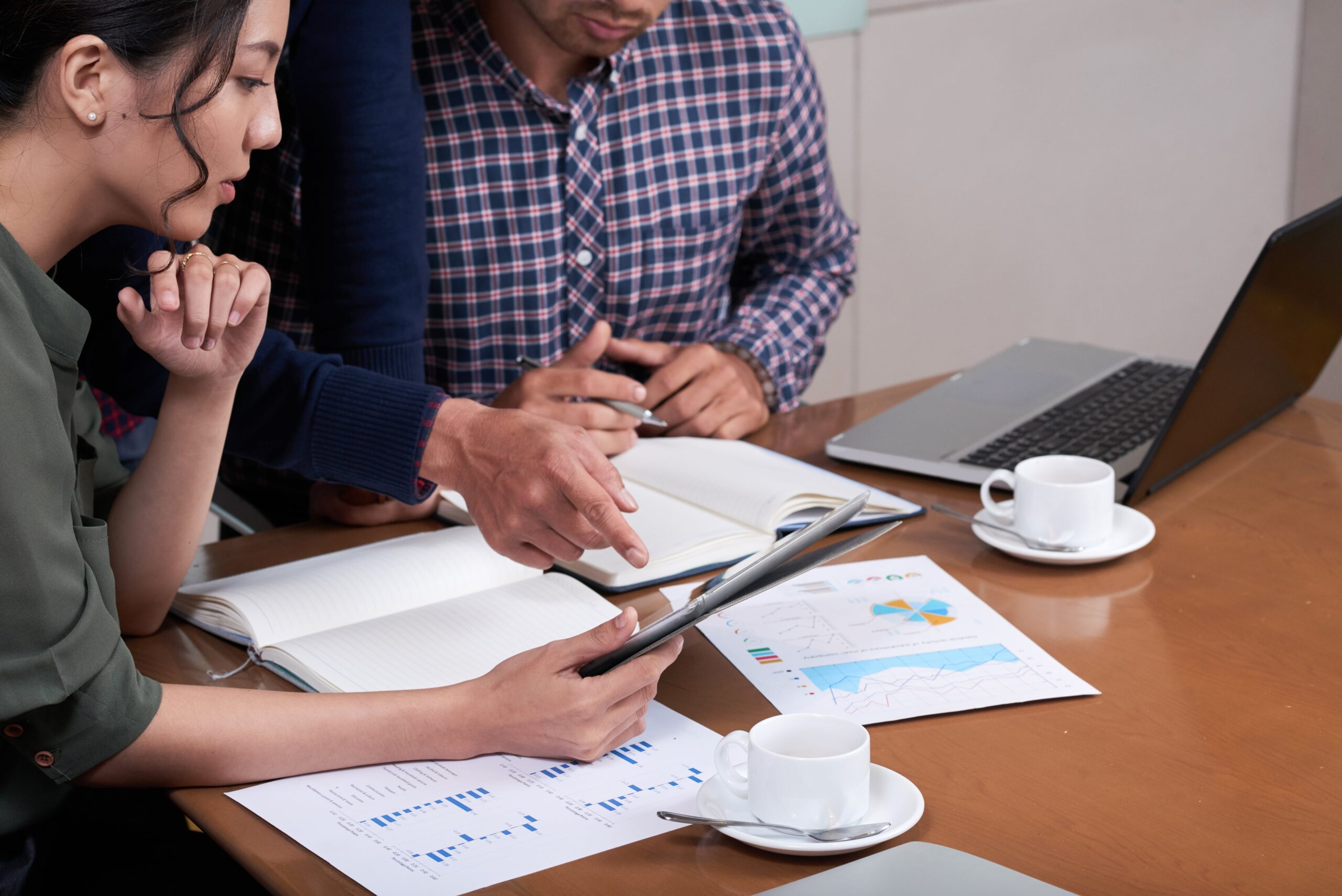 A group collaborates at a table, examining a tablet and discussing data visualizations on printed reports, with notebooks and laptops present.