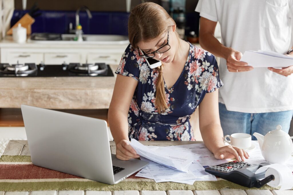 A woman reviews financial documents while talking on the phone, with a laptop and calculator on the table, indicating a busy home office setting.