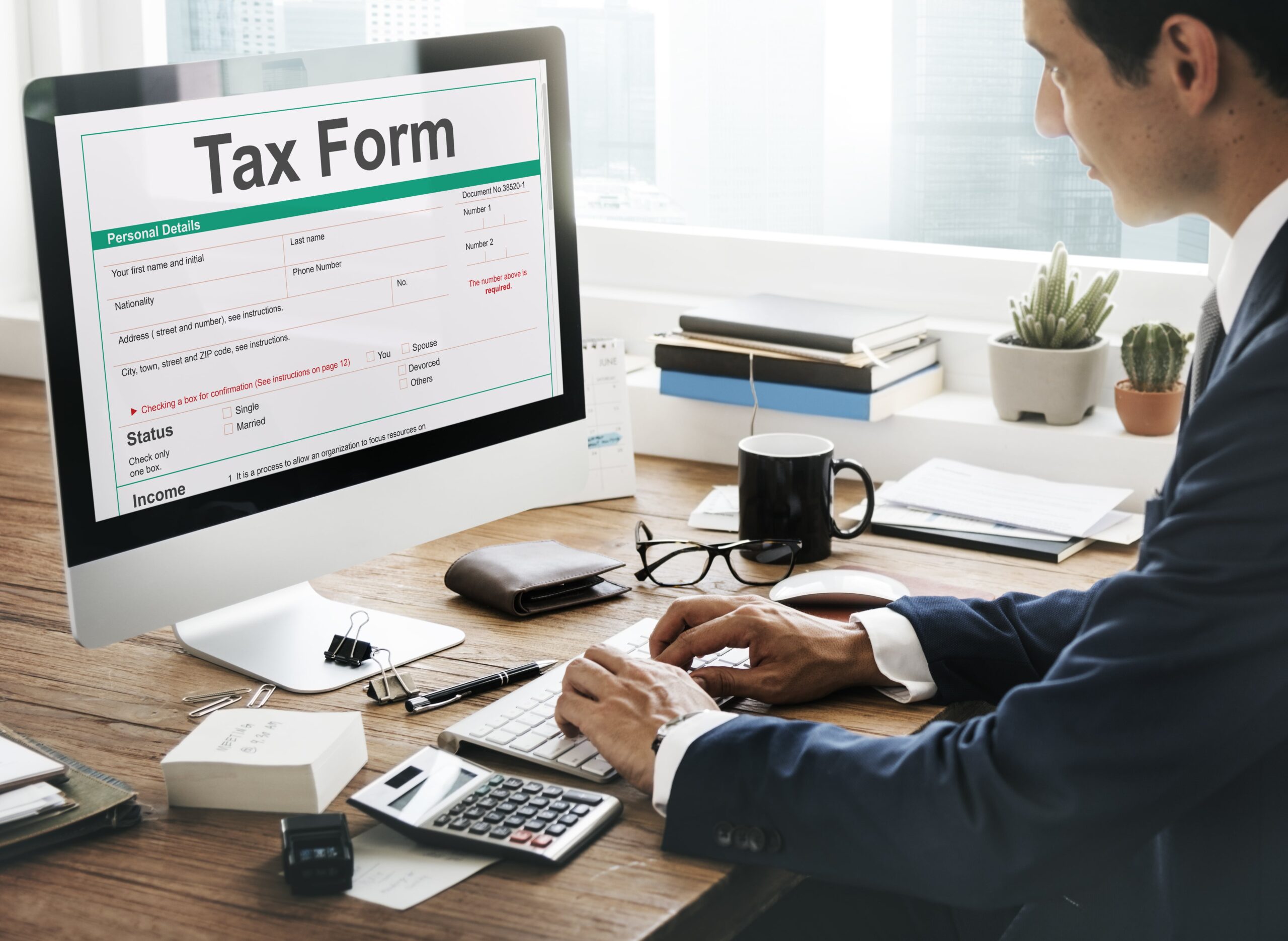 A man in a suit types on a keyboard, focusing on a tax form displayed on a desktop computer in a home office setting.