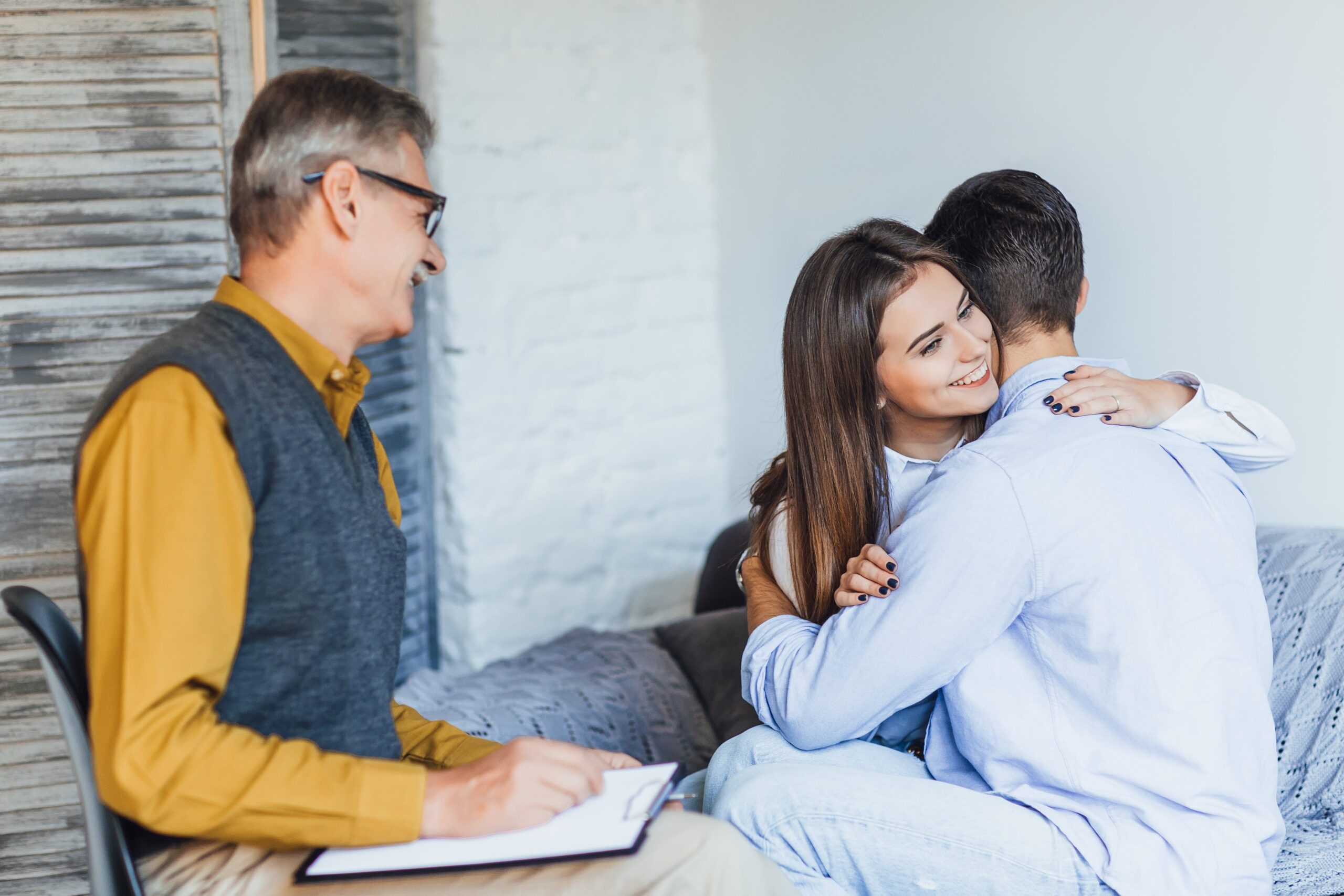 A couple embraces warmly during a therapy session, while a therapist observes and takes notes, creating a supportive environment for open communication.
