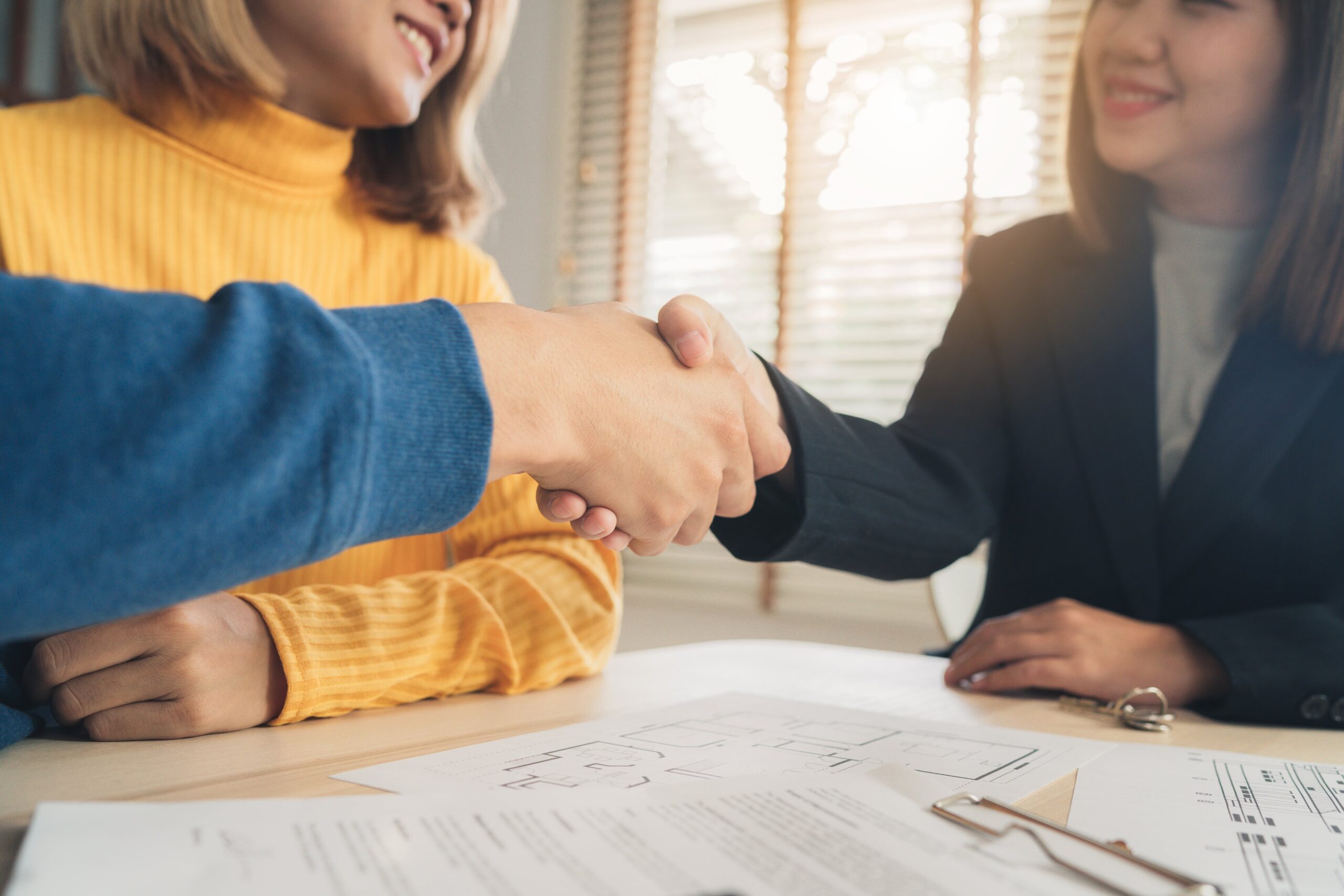 Two people shake hands across a table covered with documents, signaling a business agreement in a well-lit meeting room.