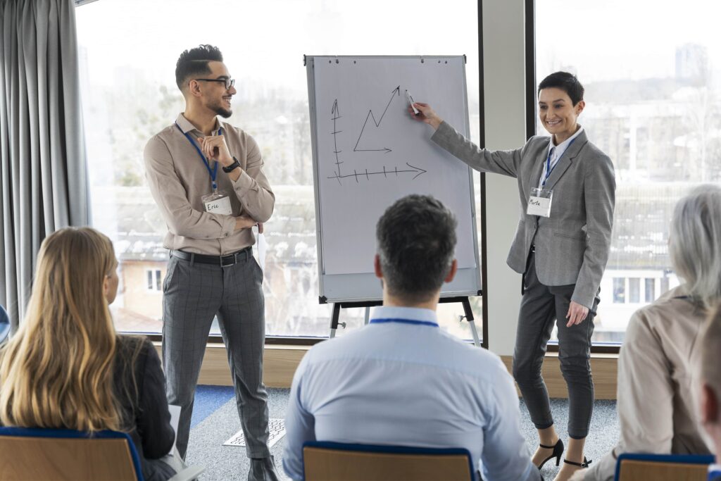 A professional presentation in a bright conference room, featuring a woman pointing at a graph on a flip chart while a man listens attentively.
