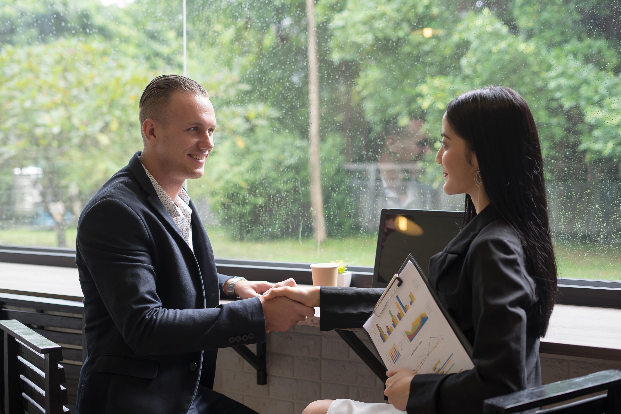 Two professionals shake hands in a café, with graphs and charts on a clipboard, symbolizing a successful business meeting amidst a rainy backdrop.