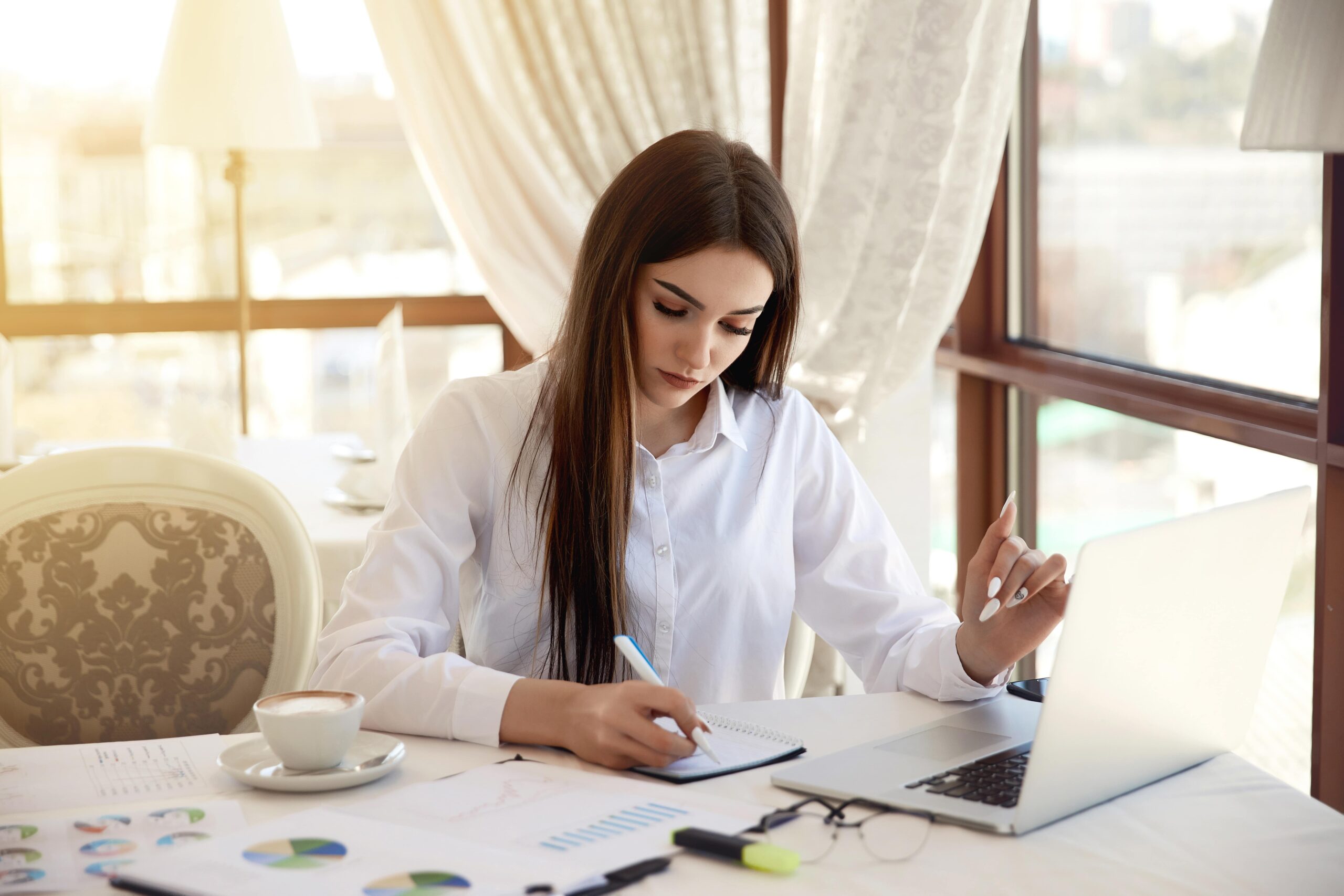A woman in a white shirt sits at a table, writing notes while working on a laptop. Charts and a coffee cup are visible, indicating a productive workspace.