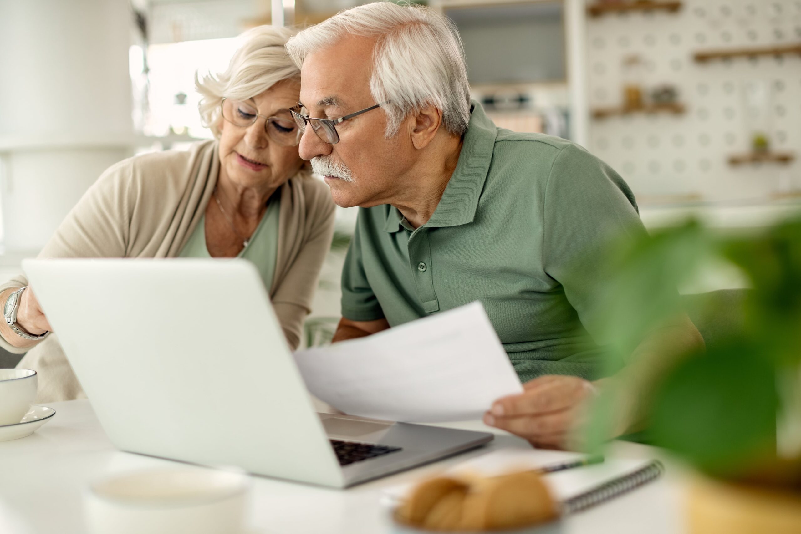 Elderly couple reviewing documents together while using a laptop in a cozy, bright kitchen, highlighting teamwork and engagement in technology.