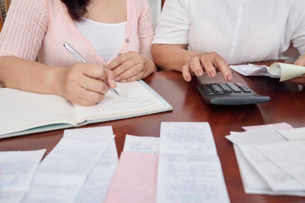 Two people review receipts and notes, one writing in a notebook while the other uses a calculator, highlighting financial organization.