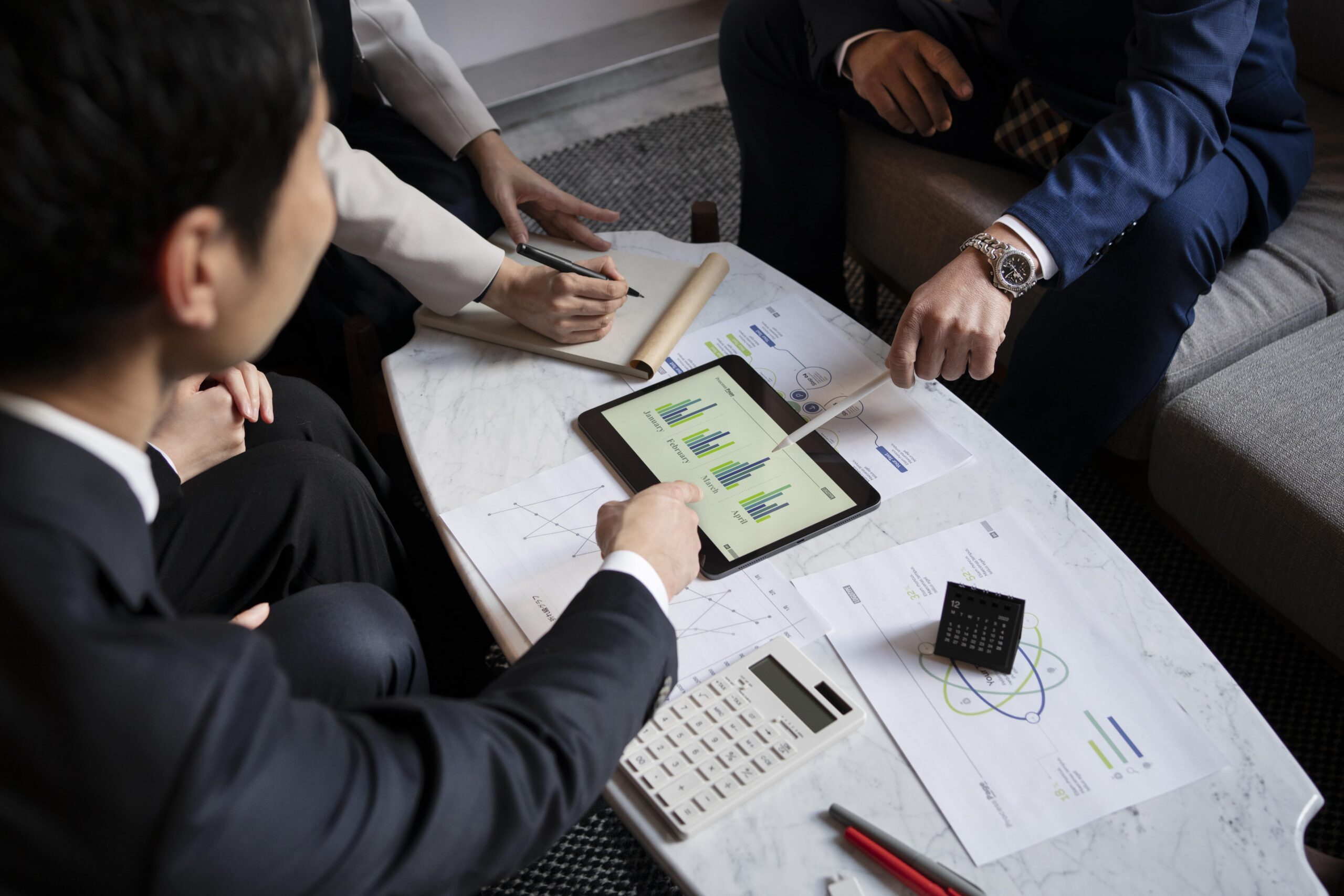Business meeting focused on analytics. Participants discuss charts on a tablet, alongside notes and a calculator on a marble table.