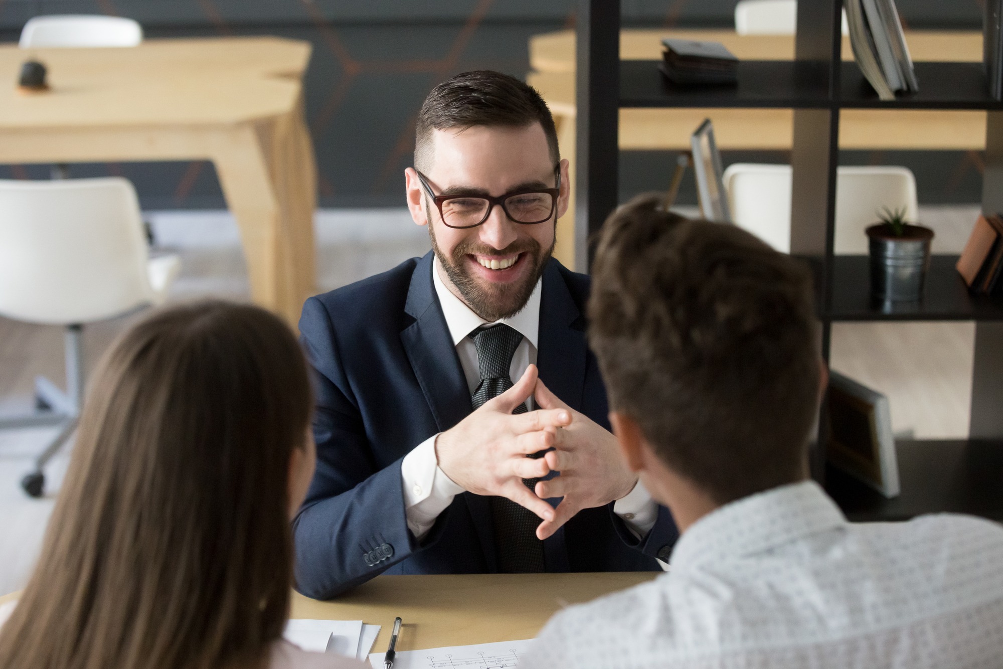 Smiling insurance agent negotiating with millennial couple in friendly atmosphere in office, realtor laughing consulting clients on property purchase, financial advisor happy with consultation meeting