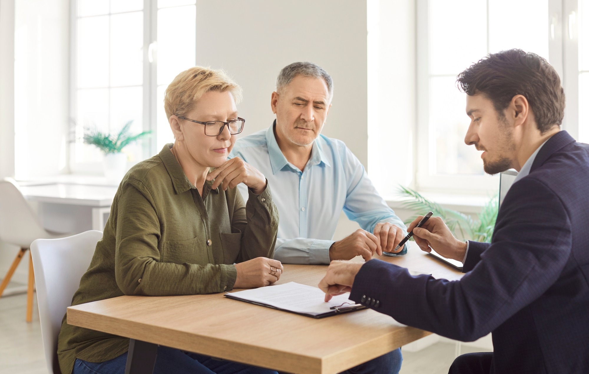 Senior couple discusses contract details with a consultant during an office meeting or negotiation. Topics include insurance, investment, and client-agent collaboration to finalize agreements.